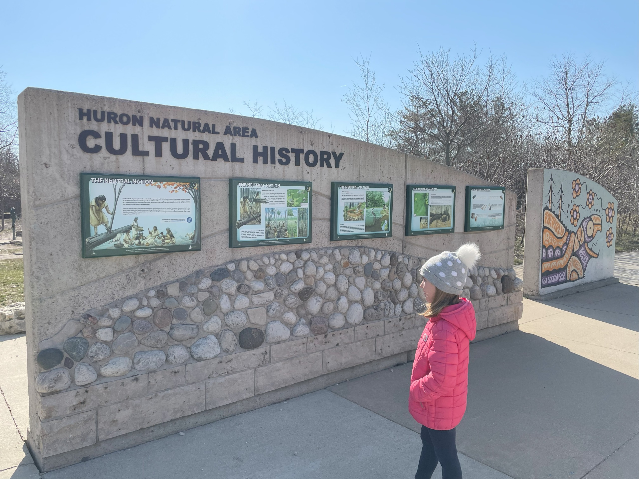 Photo of the cultural history information boards and Indigenous artwork at the entrance of Huron Natural Area. A girl in a pink coat and grey toque with a white pom-pom is looking at the boards. 