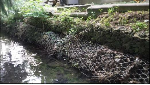 A creek lined with rock walls in a warped, slumping metal cage to form the creek bank. The bank and nearby building looks unstable due to the warped rock containment.