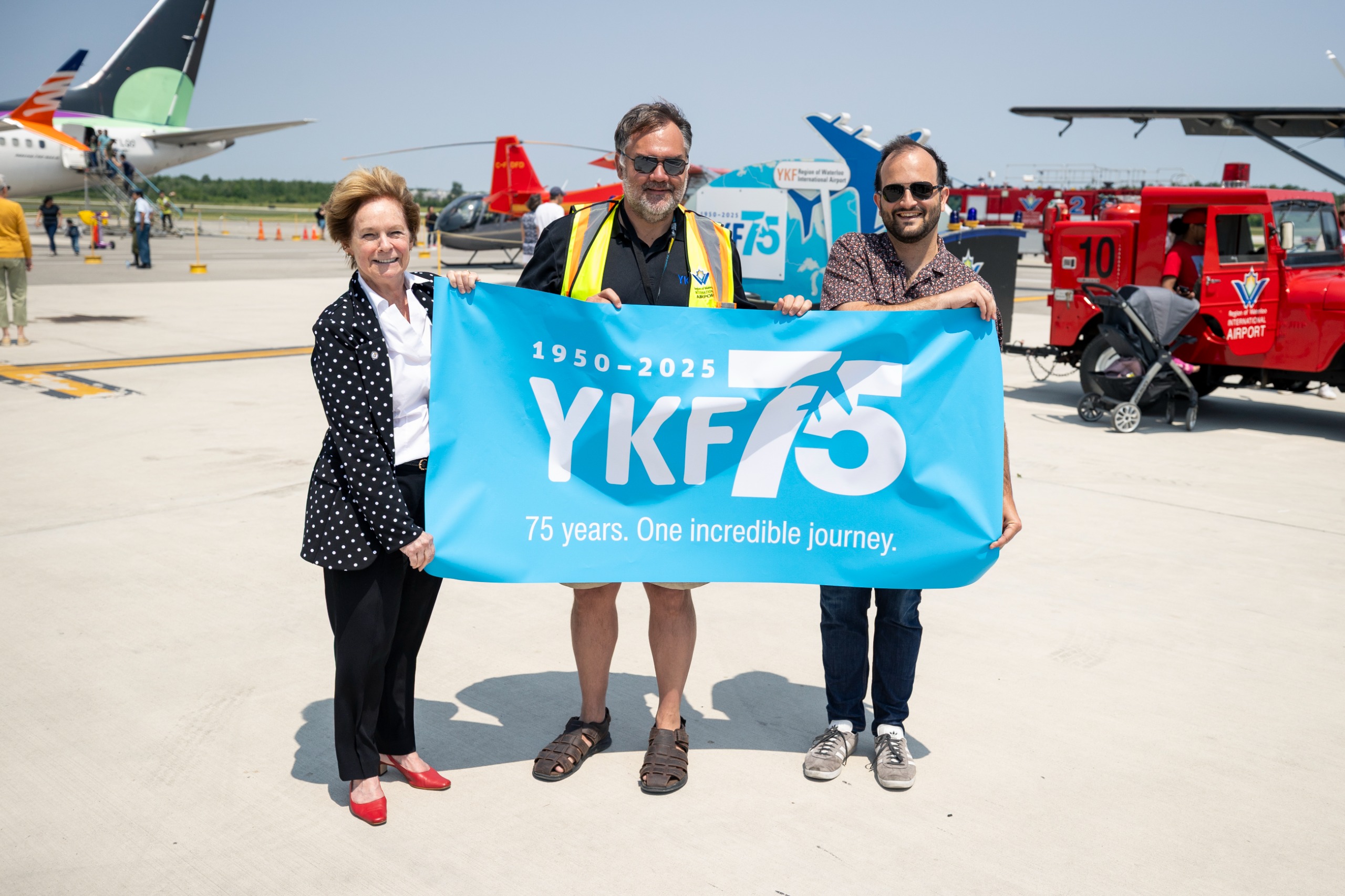 Chair Redman holds a YKF 75th Anniversary banner with Chris Wood, Director of Region of Waterloo International Airport and Councillor Matt Rodrigues 