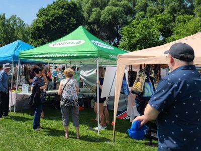 Tent with green canopy and Engage Kitchener logoat Cherry Festival. Two people are looking at the display tables underneath the tent. 