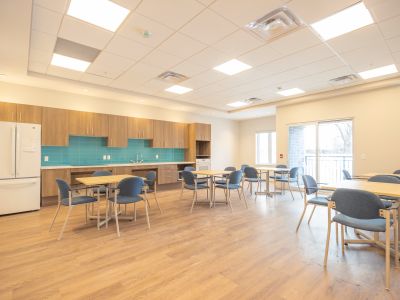 image of one of the 420 Kingscourt amenity rooms. The image shows a room with tables and chairs, a kitchen counter area with cupboards and a sink, and a patio door and window with light shining through